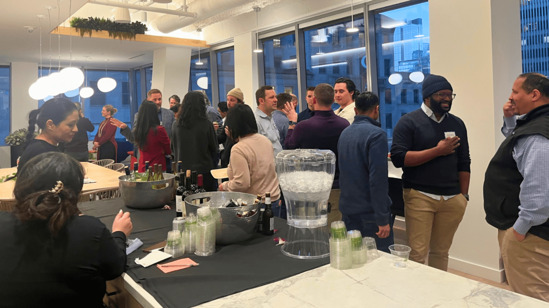 People are gathered in an indoor setting with drinks and snacks on tables, socializing at what appears to be a casual event.